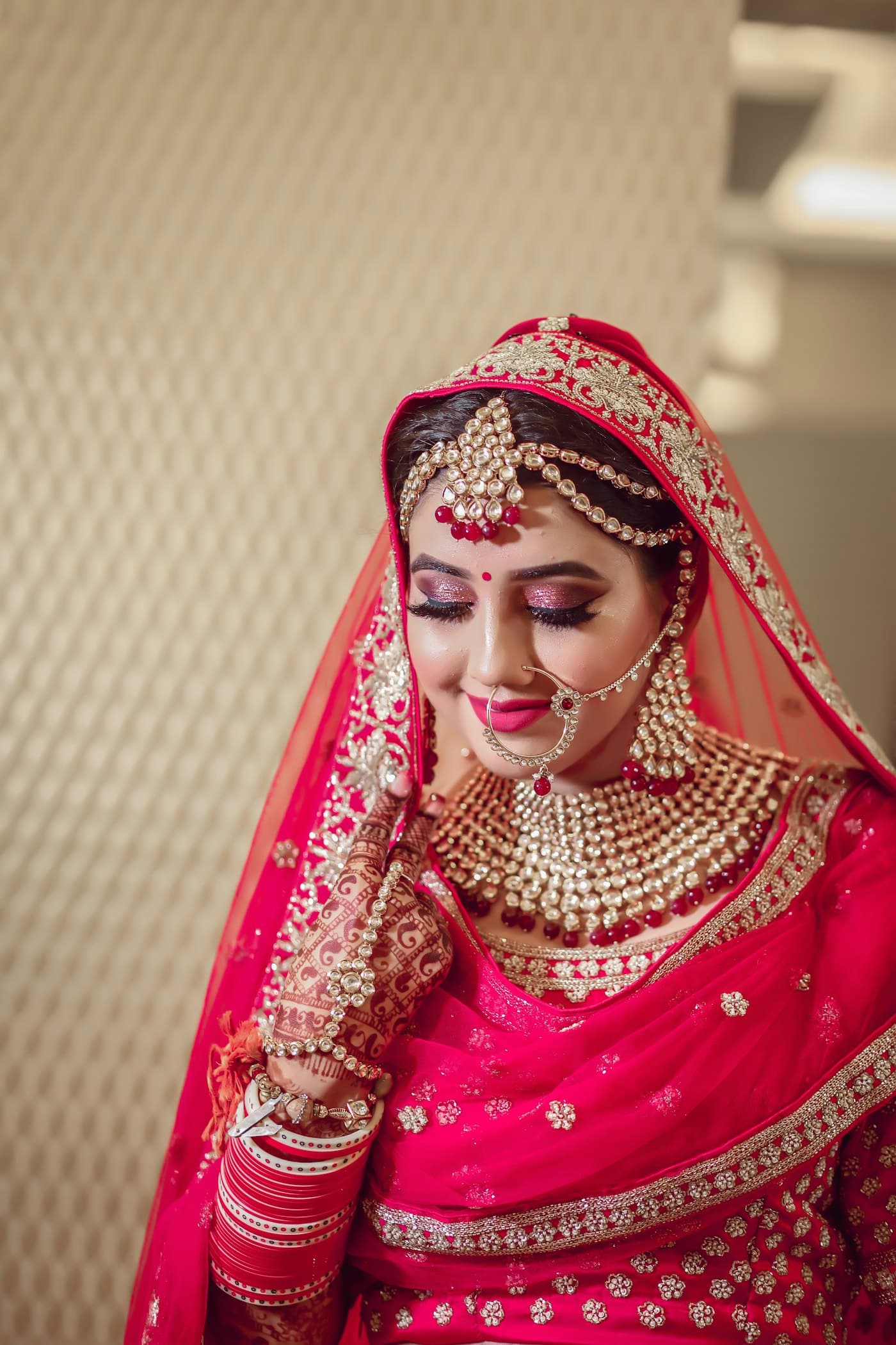 A bride in a red and gold lehenga looks down, with mehendi visible on her hand.