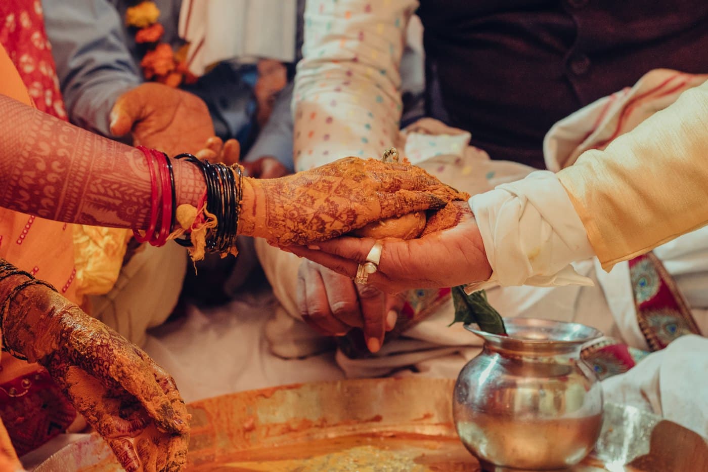 Hands covered in turmeric and mehendi during a wedding ritual, beside a silver pot.