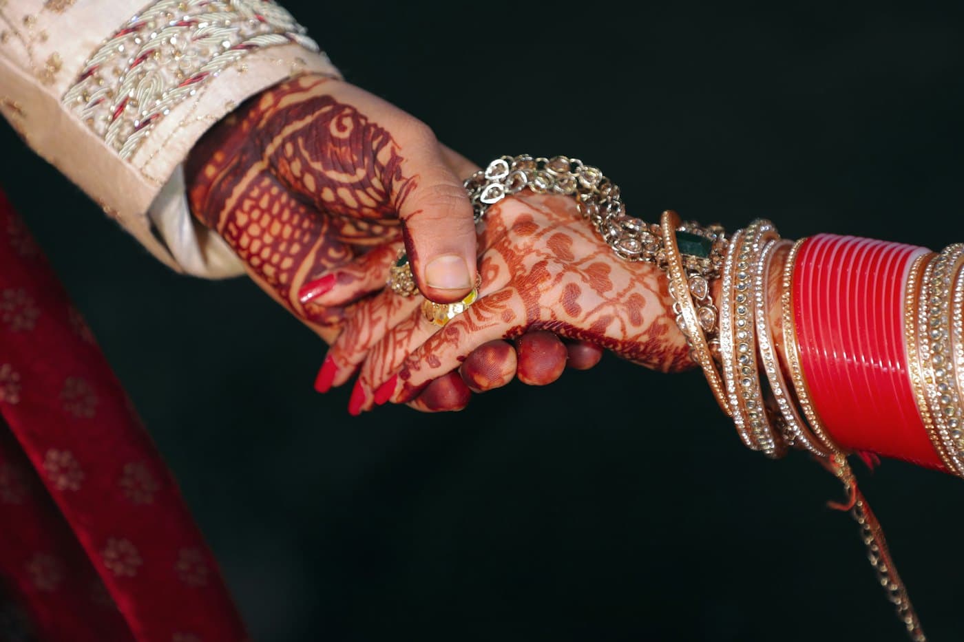 A bride and groom hold hands, showing mehendi, bangles, and embroidered sleeves.