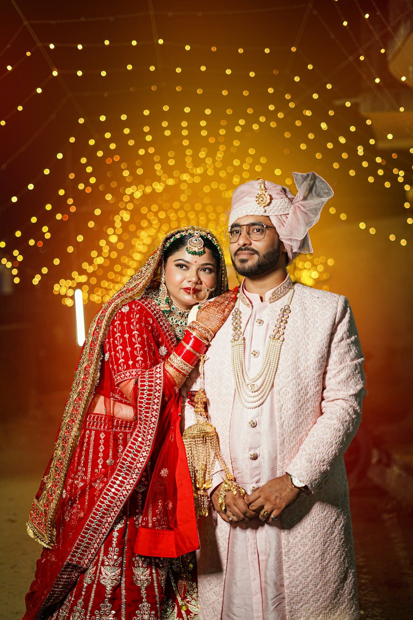 A bride and groom in embroidered wedding clothes pose under warm evening lights.